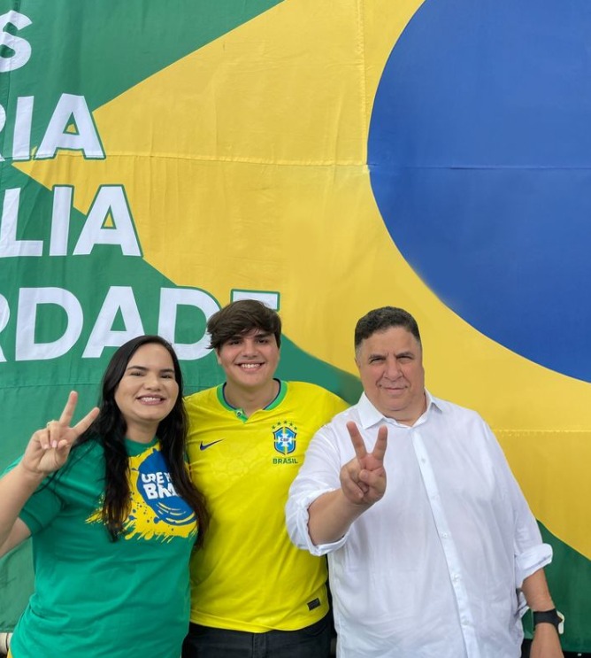 Foto de três pessoas em frente a uma grande bandeira do Brasil. Uma delas é Michele Collins, mulher branca de cabelos escuros lisos, vestindo uma camiseta verde com “Brasil” escrito em amarelo e fazendo um sinal de paz com a mão. A pessoa do meio é seu filho Alef, um jovem branco, de cabelos lisos, que está usando uma camisa da seleção brasileira de futebol, na cor amarela. À direita, Cleiton Collins, homem branco de meia-idade, veste uma camisa branca e também faz o sinal de paz, com o rosto parcialmente escondido. Todos os três estão, visivelmente, acima do peso.