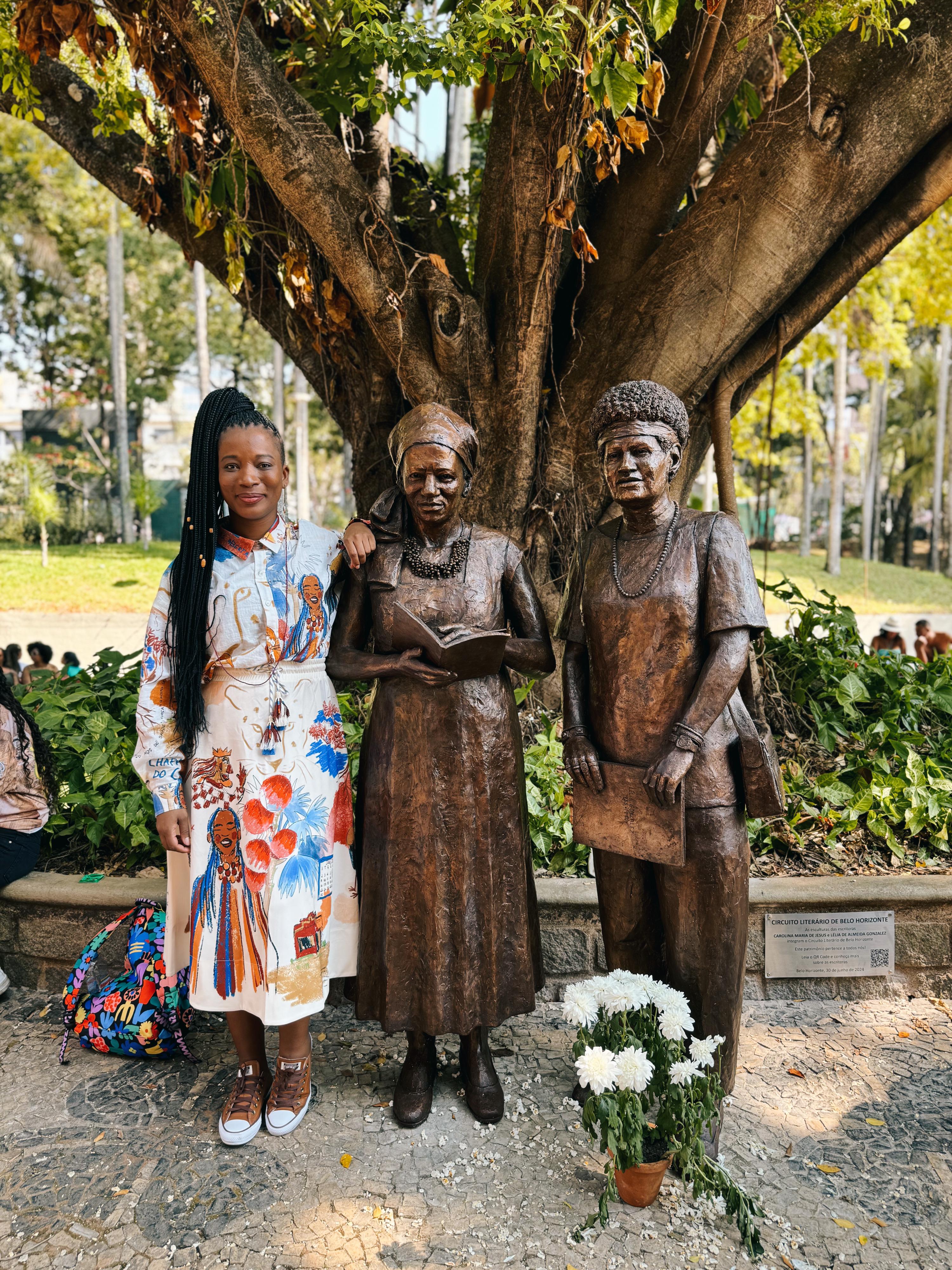 Foto de Etiene Martins ao lado das estátuas de Carolina Maria de Jesus e Lélia Gonzalez, esculpidas na cor marrom e instaladas em um parque público, cercadas de plantas e árvores.