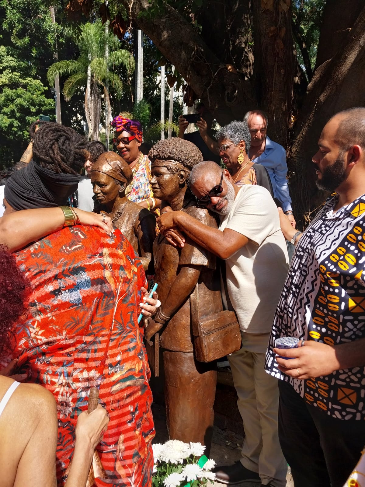 Foto de várias pessoas negras, com roupas coloridas, em torno de duas estátuas de mulheres negras esculpidas na cor marrom. No centro da imagem, um homem negro, idoso, de barba branca e óculos escuro, abraça uma das esculturas.