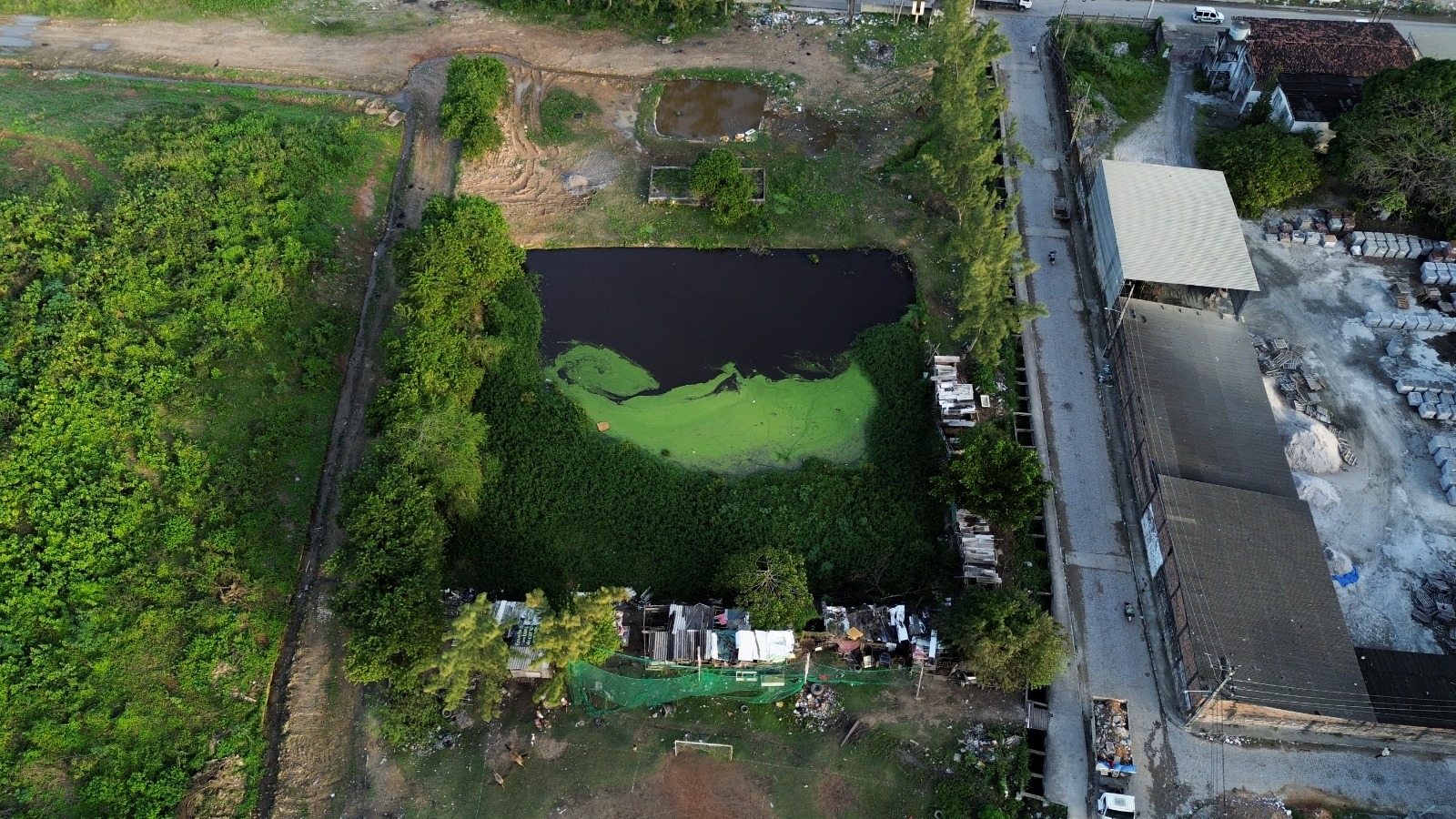 Foto aérea da lagoa de chorume de Aguazinha. A imagem é uma vista aérea de uma paisagem que inclui um grande corpo d’água escuro com algas verdes ou matéria vegetal em sua superfície. Ao redor do corpo d’água, há áreas de vegetação densa e um caminho de terra, além de algumas áreas de terra nua. À direita do corpo d’água, há um grande prédio com telhado cinza, e ao lado dele, pilhas de materiais que parecem ser para construção ou uso industrial. Também há estruturas menores com telhados de várias cores espalhadas na borda inferior do corpo d’água. O canto superior esquerdo mostra outra área aberta com o que parece ser água parada e mais vegetação.