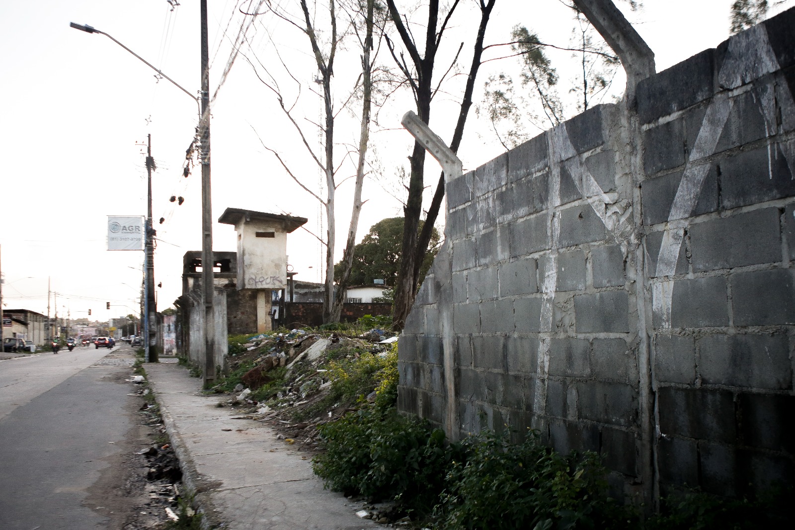 Foto do lado externo do muro do lixão de Aguazinha. À direita, há um alto muro de concreto cinza, desgastado e sem pintura. À esquerda do muro, há uma área com vegetação exuberante, com várias plantas verdes e arbustos. Mais à esquerda, há uma rua com postes de utilidade pública e fios suspensos. Ao longo dessa rua, há edifícios que parecem estar em diferentes estados de deterioração ou construção, com um deles mostrando tijolos expostos e outro tendo uma pequena estrutura semelhante a uma torre no topo. O céu está nublado, sugerindo que pode ser início da noite ou um dia nublado. Não há pessoas visíveis nem veículos em movimento na cena.