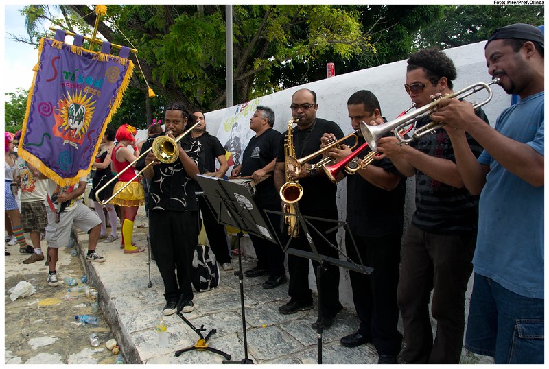 A foto retrata um grupo de músicos se apresentando ao ar livre em uma rua durante o carnaval de Olinda. Eles seguram instrumentos de sopro, como trombones e trompetes. Uma pessoa carrega uma grande faixa com cores vibrantes e padrões decorativos. Todos os músicos são homens e usam roupas pretas, porém estão vestidos informalmente.