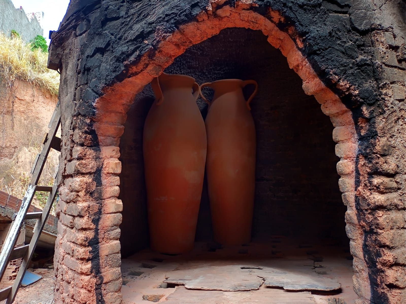 A foto mostra um forno de tijolos tradicional com uma abertura em arco. Dentro do forno, há dois grandes potes de terracota sem esmalte, posicionados verticalmente um ao lado do outro. Os potes parecem ter sido recentemente feitos e provavelmente estão no forno para serem queimados, um processo que endurece objetos de argila por meio de altas temperaturas. O interior do forno está escurecido, possivelmente devido a queimas anteriores, e contrasta com a cor laranja brilhante dos potes. O fundo fora do forno está superexposto devido às condições de iluminação intensa, tornando os detalhes indiscerníveis.