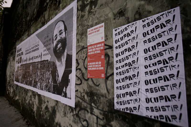 A imagem mostra uma parede com vários cartazes. À esquerda, há um grande cartaz preto e branco de uma pessoa com barba e bigode, vestindo terno e gravata. A pessoa está sorrindo, e abaixo da imagem há um texto que não está totalmente visível devido ao ângulo da fotografia. À direita, há vários cartazes menores vermelhos com texto branco em negrito repetindo as palavras “OCUPAR,” “RESISTIR,” em letras maiúsculas. Esses cartazes estão dispostos em duas colunas; alguns estão diretamente na parede, enquanto outros se sobrepõem aos já existentes. A parede de fundo parece envelhecida, com desgaste visível e algumas marcas de grafite.