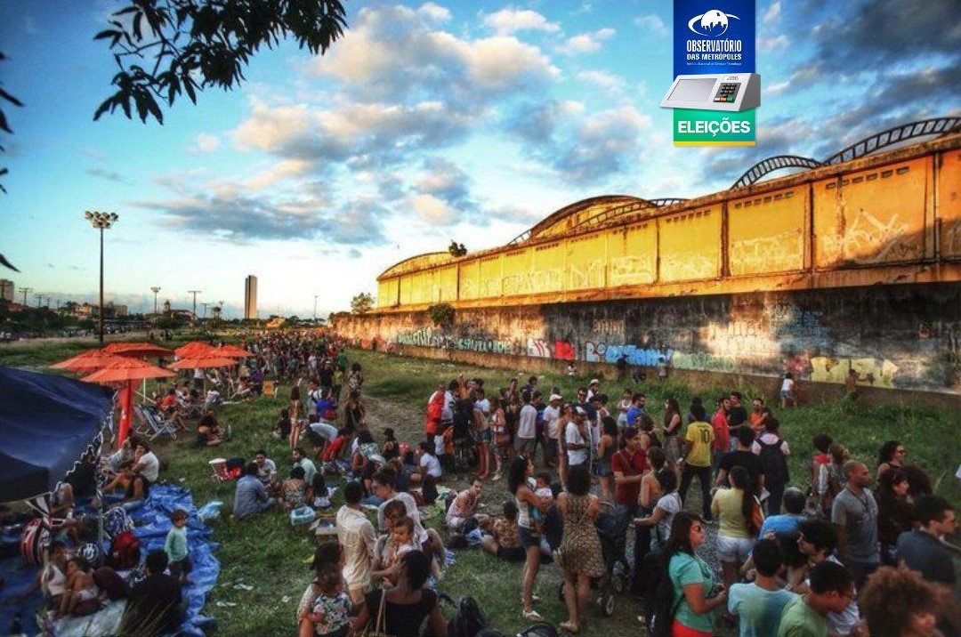 A imagem mostra uma grande reunião de pessoas ao ar livre, provavelmente no final da tarde, devido à iluminação quente. O cenário é uma área urbana, e a multidão está concentrada em um campo gramado, com algumas pessoas em pé e outras sentadas. Ao fundo, há estruturas longas e amarelas que se assemelham a vagões de trem ou armazéns, decoradas com grafites. Acima dessas estruturas, o céu está parcialmente nublado, com algumas áreas azuis visíveis. No lado direito da imagem, há uma estrutura azul em forma de tenda, proporcionando sombra a uma área embaixo dela. No canto superior direito da foto, há um logotipo com o texto “Observatório das Metrópoles” e abaixo “Eleições,” sugerindo que este evento pode estar relacionado a uma eleição ou reunião política.