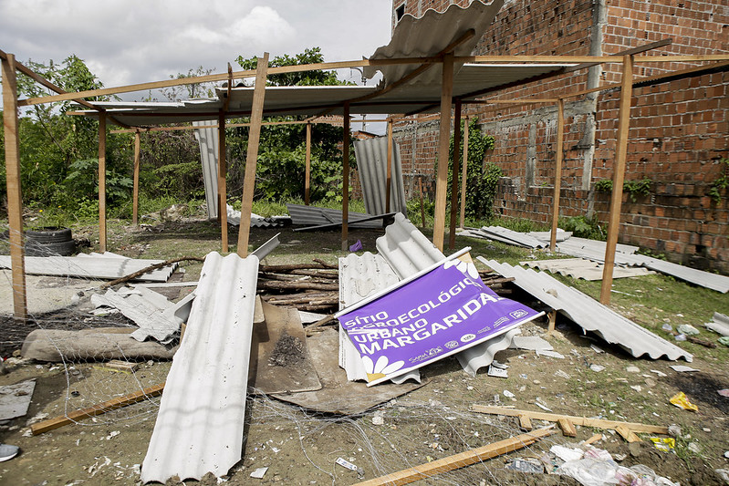 A foto retrata uma cena de destruição na horta do Jiquiá. Há uma estrutura parcialmente colapsada com vigas de madeira expostas e um telhado de metal ondulado, parte do qual caiu ao chão. A área circundante está repleta de destroços, incluindo pedaços quebrados do telhado de metal, tijolos e outros escombros não identificáveis. Um grande banner roxo com texto em amarelo e branco sinalizando o Sítio agroecológico urbano margaridas está entre os destroços.