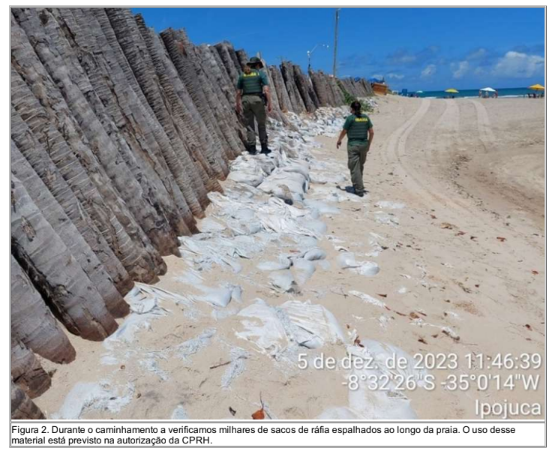 A foto retrata uma cena de praia, onde uma longa fileira de grandes sacos de areia branca está disposta ao longo da costa, paralela a um alto muro de contenção de madeira. O céu está limpo, com poucas nuvens, e a areia é de cor clara. Duas pessoas em uniformes verdes caminham ao longo dos sacos de areia, inspecionando-os. A foto inclui um carimbo de data e hora no canto inferior direito, indicando que foi tirada em 5 de dezembro de 2023, e também apresenta coordenadas e o nome do local “Ipojuca”.