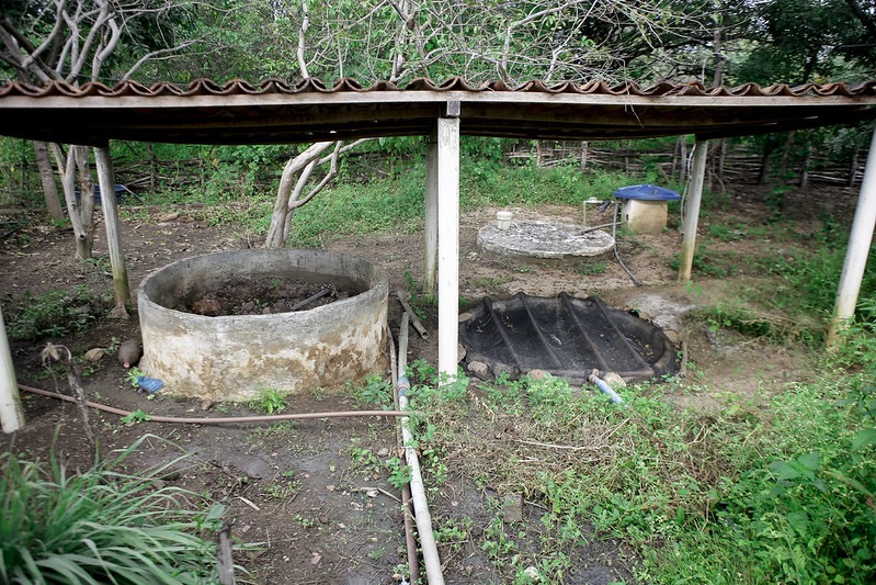 A foto retrata uma cena rural ao ar livre com vários elementos que sugerem um ambiente de agricultura ou cultivo. Na frente, há uma grande estrutura circular de concreto, semelhante a um poço ou cisterna, que parece estar desgastada e sem uso, com detritos em seu interior. Ao lado dessa estrutura, há uma área coberta com pilares brancos e um telhado de metal corrugado. À direita da estrutura de concreto, há uma lona de plástico preto no chão, possivelmente indicando uma área para compostagem ou outra atividade agrícola. O fundo mostra vegetação densa e parte de uma cerca de madeira que delimita a área