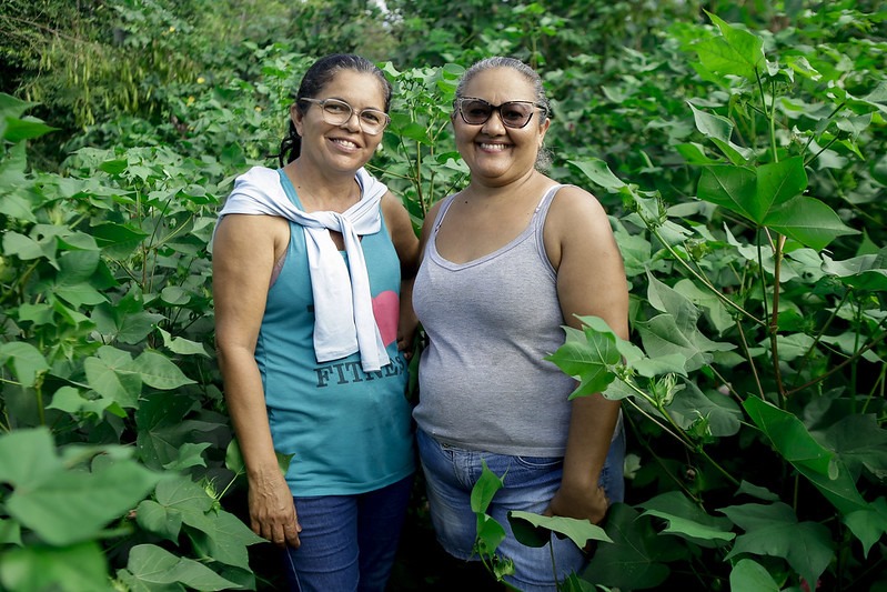 Na imagem, vemos duas mulheres em meio a uma vegetação densa e verde. A mulher à esquerda – negra, de cabelos lisos e presos de meia-idade, mas com sorriso jovial e óculos - está usando uma camiseta regata azul com um desenho rosa e uma palavra inelegível com letras pretas, com uma blusa ou casaco cinza claro sobre os ombros e amarrado no pescoço. A mulher da direita veste uma blusa cinza sem mangas. Ela é também tem a pele escura, cabelos grisalhos presos e usa óculos escuros. O cenário é exuberante e verde, sugerindo um ambiente natural, possivelmente tropical.