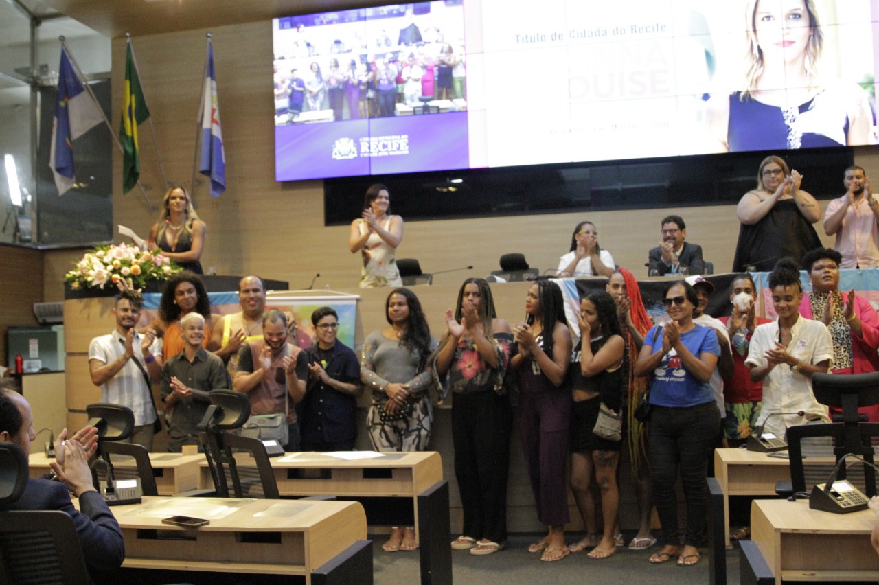 Foto do plenário da Câmara Municipal do Recife, com várias pessoas vestindo roupas casuais perfiladas junto à mesa diretora. Algumas estão aplaudindo, enquanto, no alto à esquerda, Dayanne Louise, mulher loira, de cabelos ondulados e pele bronzeada, vestindo camiseta preta decotada. Ao fundo, painel exibe imagens do que está acontecendo no plenário e foto em close da homenageada.