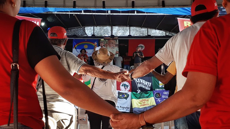 A imagem mostra um grupo de pessoas reunidas em um evento público ou manifestação. No centro, dois pares de mãos estão unidas, umas de pessoas que estão de costas para a câmera, à esquerda, e outras da direita, simbolizando união ou solidariedade. Ao fundo, há outros participantes e banners com símbolos do MST e textos, indicando que pode ser um encontro político ou social. Um dos banners tem a ilustração de uma pessoa usando um chapéu vermelho e segurando o que parece ser uma bandeira.