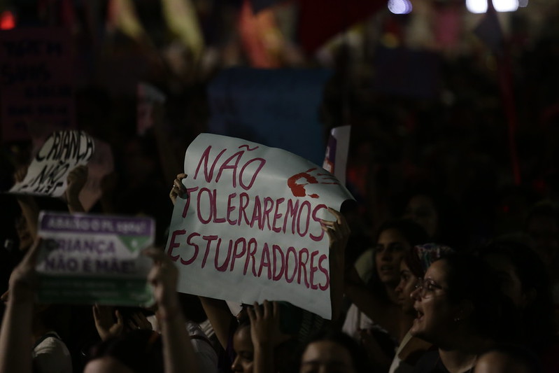 A imagem mostra uma cena de protesto à noite. Há várias pessoas segurando cartazes, com um cartaz em primeiro plano que é mais visível que os outros. Este cartaz tem uma mensagem escrita em português: “Não toleraremos estupradores.” A iluminação é fraca e os rostos das pessoas não estão claramente visíveis. A presença de vários cartazes sugere que esta é uma manifestação por uma causa social, especificamente contra a violência sexual.