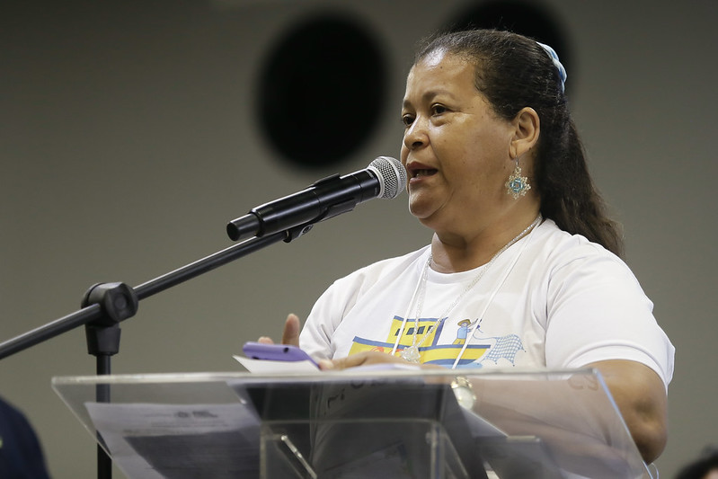 Foto da pescadora Maria Aparecida de Santana falando ao microfone. Ela é uma mulher negra, de meia idade, vestindo uma camiseta branca com um desenho de um barco amarelo e azul na parte da frente da vestimenta, Ela usa brincos grandes em forma de estrela e tem os cabelos presos em coque por trás do pescoço.