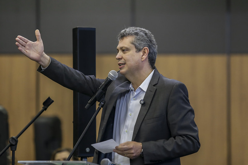 Foto do ministro Márcio Macedo, homem branco, maduro, de cabelos pretos com fios brancos. Ele está falando ao micrfone, gesticulando com a mão direita, em um ambiente interno de paredes marrons, usando um paletó cinza e camisa branca.