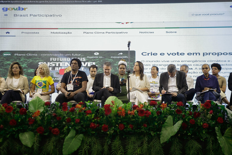 A imagem mostra um grupo de pessoas sentadas em frente a um palco com um fundo que exibe o logo e o nome “Brasil Participativo” junto com alguns textos em português. No centro da foto, está a governadora Raquel Lyra., à direita está a ministra do Meio Ambiente Marina Silva, com um homem de paletó cinza e cabeça abaixada entre as duas. Na frente delas, há uma fileira de plantas em vasos com folhagem verde e flores vermelhas vibrantes