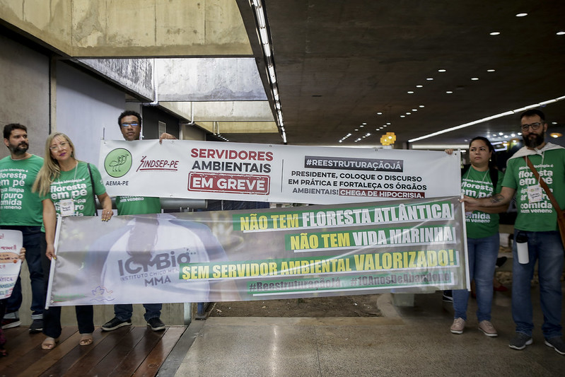 Foto de cinco pessoas, sendo três homens e duas mulheres, usando camisetas verdes onde é possível ler floresta em pé sobre outras palavras que não são legíveis. Esse gupo segura duas faixas, uma sobre a outra. A faixa de cima é branca com letras vernelhas e pretas formando o seguinte texto: Servidores ambientais em greve - reestrutruração já - presidente, coloque o discurso em prática e fortaleça os órgãos ambientais! Chega de hipocrisia. A outra faixa tem frases copm letras amarelas e brancas com fundo verde formando o seguinte texto: Não tem floresta atlântica! Não tem vida marinha! Sem servidor ambiental valorizado!