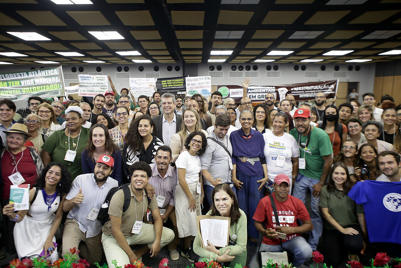 Foto de dezenas de pessoas, homens e mulheres em um grande salão. Elas estão usando roupas variadas, tendo vários cartazes nas cores pretas e brancas ao fundo.