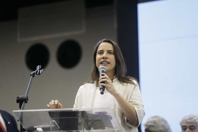 Foto de Raquel Lyra segurando um microfone junto a uma mesa de material transparente. Ela está usando um cardigã bege sobrepondo uma camisa branca.