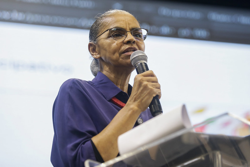 Foto da ministra Marina Silva. Mulher negra, idosa, de óculos e cabelos presos em coque atrás do pescoço. Ela está usando blusa azul escuro e está segurando o microfone enquanto fala em público junto a uma mesa de material transparente com folhas de papel em cima.