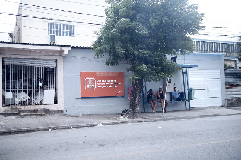 A foto mostra uma cena urbana durante o dia. Há um prédio com fachada azul e barras de segurança brancas nas janelas. Na parede deste prédio, há uma grande placa laranja com texto branco e amarelo anunciando uma “Creche-Escola” chamada “Nossa Senhora das Graças - Matriz”. Em frente ao prédio, há um ponto de ônibus onde duas pessoas estão sentadas, esperando. Ao lado do ponto de ônibus, há uma árvore verde e frondosa que parcialmente obscurece a visão de uma das pessoas e do prédio.