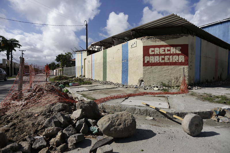 A imagem mostra uma cena ao ar livre de um local de construção ou demolição. Há um prédio com paredes pintadas de azul claro e bege, com as palavras “CRECHE PARCEIRA” escritas em vermelho na seção bege. O céu está parcialmente nublado, e há cercas de metal parcialmente cobertas com uma malha plástica laranja, indicando acesso restrito à área. Entulhos estão espalhados pelo primeiro plano, incluindo pedaços de concreto e vergalhões. Um martelo de demolição está no chão, próximo ao centro da imagem.