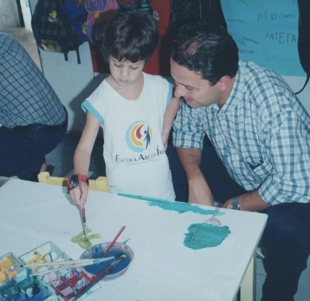 Foto envelhecida de Eduardo Campos, de camisa quadriculada, agachado, ao lado de seu filho João junto a uma banca escolar.