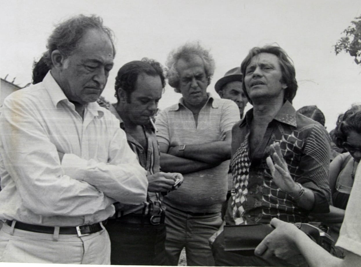 A foto em preto e branco mostra um grupo de cinco homens em pé, próximos uns dos outros. São Miguel Arraes, um homem calvo ao centro, Jarbas Vasconcelos e Marcos Freire. Eles parecem estar envolvidos em uma conversa ou reunião, com algumas pessoas de braços cruzados ou mãos juntas, sugerindo discussão ou espera. O cenário parece ser ao ar livre, com árvores e um céu aberto ao fundo.