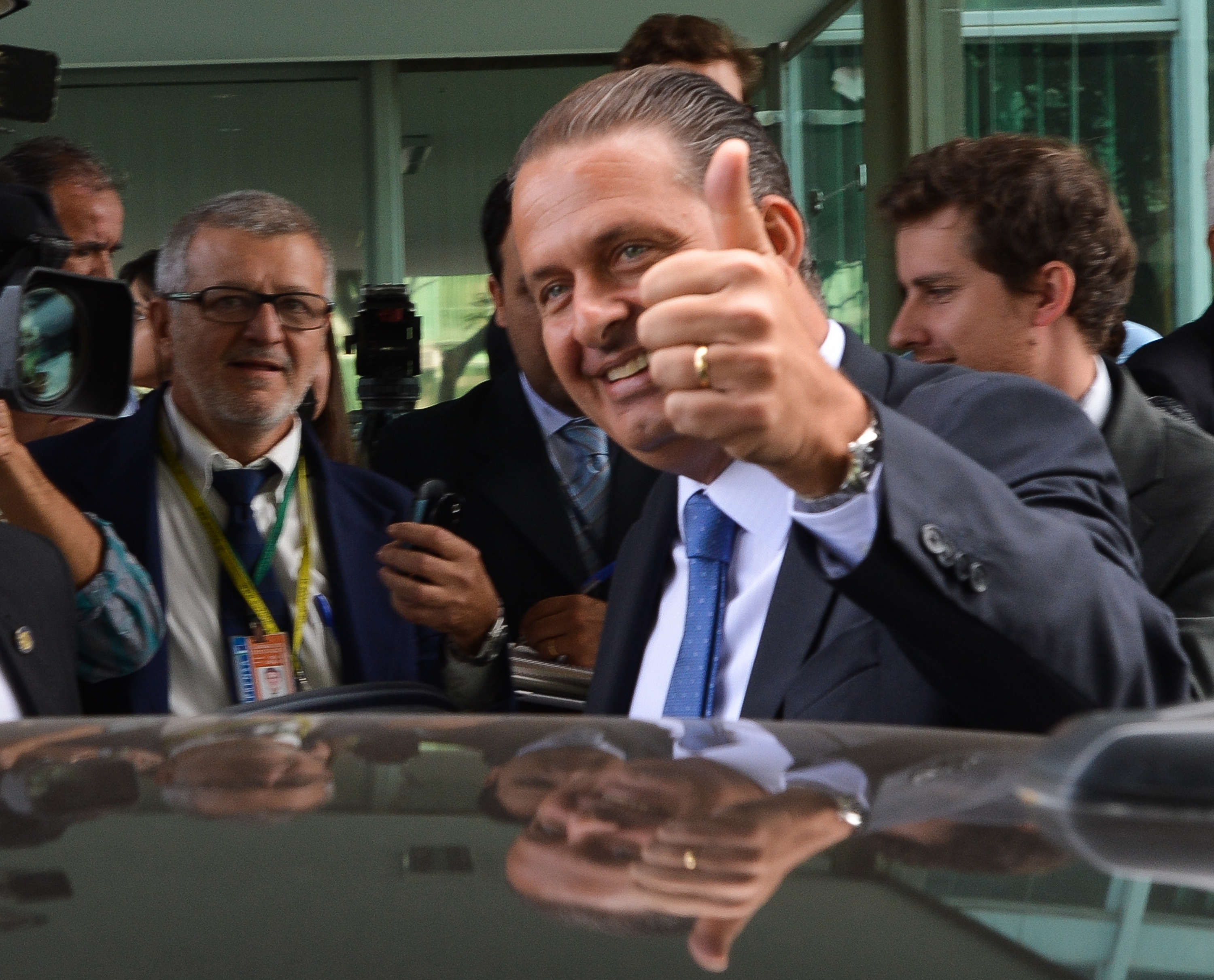 Foto de Eduardo Campos fazendo gesto de legal, com o polegar voltado para cima, junto a um carro em cujo capô se reflete sua imagem. Ele parece estar chegando a um local, cercado de outros homens que o esperam.