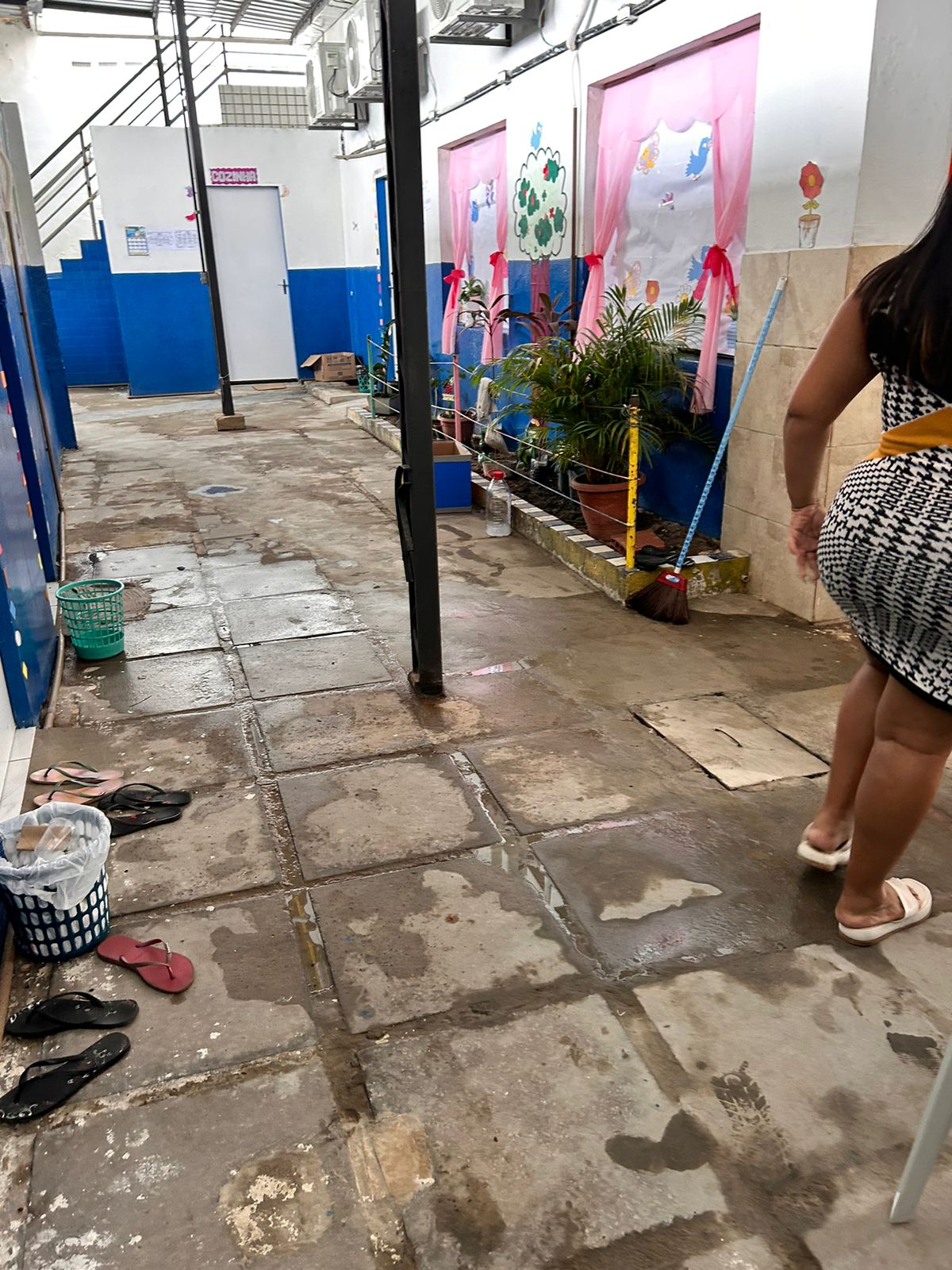 A imagem mostra um corredor interno que parece ser de uma escola ou uma instalação educacional similar. O chão é de lajotas de cimento, com algumas áreas mostrando poças de água e sinais de desgaste. À esquerda, há paredes azuis e uma série de colunas que sustentam a estrutura. À direita, há pôsteres ou obras de arte coloridas exibidas em paredes rosas atrás de janelas de vidro, decoradas com arte infantil envolvendo árvores e figuras. Plantas em vasos podem ser vistas na frente dessas janelas, adornadas com fitas vermelhas. Também há um balde verde perto de uma das plantas à esquerda. No primeiro plano, parte de uma pessoa vestindo um vestido preto e branco com padrão é visível na borda direita da imagem; apenas da cintura até a metade da coxa pode ser visto. Outro par de calçados está abandonado no chão ao lado dessa pessoa.