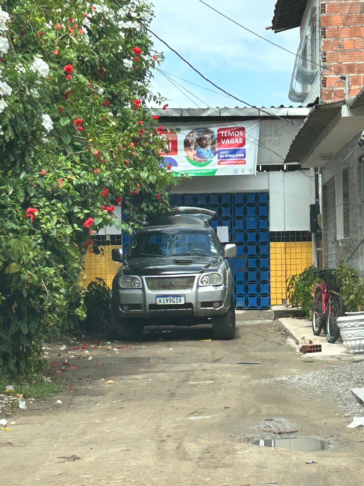 A imagem mostra uma cena urbana, especificamente uma rua ou entrada estreita. No centro, há um carro prateado estacionado, com a frente voltada para a câmera. O carro está parcialmente sobre a calçada e parcialmente na rua. Ao redor do carro, há elementos que sugerem ser uma área residencial: à esquerda, há um muro amarelo com folhagem verde pendendo sobre ele, e à direita, parece haver um portão aberto que leva a um prédio com paredes azuis e brancas. Acima dessa entrada, há uma placa com texto e imagens de crianças. Atrás do carro e acima da linha do telhado dos prédios próximos, fios elétricos são visíveis contra um céu claro.