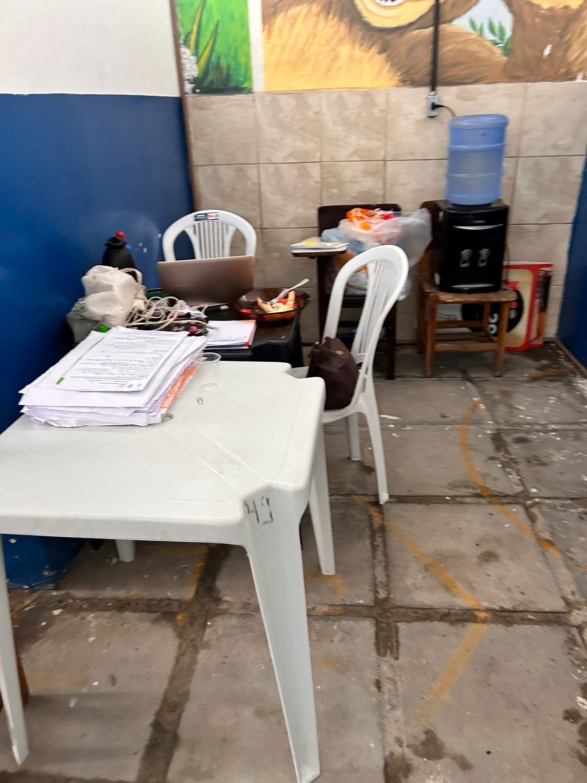 A foto mostra um ambiente interno que parece ser um espaço de trabalho ou estudo casual. Há uma mesa de plástico branca em primeiro plano com vários itens sobre ela, incluindo papéis, livros e o que parece ser um par de óculos. À direita dessa mesa, há outra cadeira de plástico branca voltada para longe da câmera. Ao fundo, há outra mesa cheia de itens diversos e uma bolsa preta sobre uma das cadeiras. No canto direito, há um dispensador de água sobre uma estrutura vermelha com copos disponíveis para uso. O chão é de lajotas de cimento cinzentas, e parte de uma parede azul é visível com o que parece ser uma imagem ou pintura.