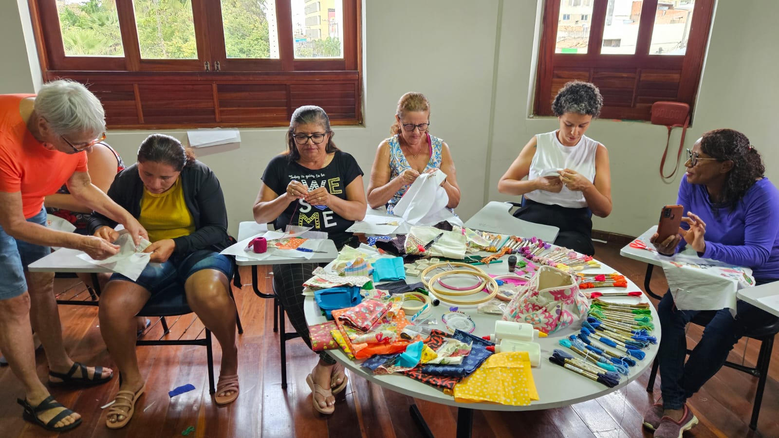 Foto de seis mulheres de meia idade, das quais cinco estão sentadas enquanto fazem bordados, com algumas peças coloridas desse tipo de artesanato dispostas sobre uma mesa redonda. A sexta mulher, usando uma blusa laranja, está em pé, à esquerda da foto, curvada sobre uma das mulheres sentadas como se estivesse ajudando no trabalho artesanal.