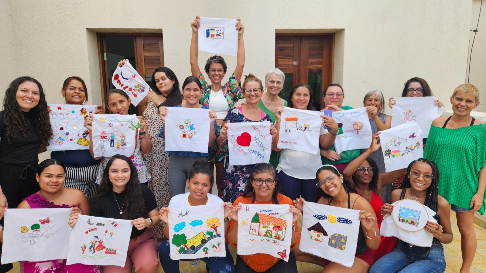 Foto de 20 mulheres reunidas em duas fileiras (13 delas estão em pé e set agachadas), exibindo peças de artesanato colorido, com imagens variadas, no formato quadrado. Elas estão diante da fachada de um imóvel com janelas e porta marrons.