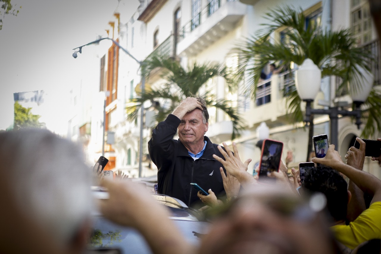 Foto de Jair Bolsonaro com semblante tenso e mão direita na cabeça. Ele está acima de uma multidão da qual só se vê mãos desfocadas e telefones celulares colocados acima das cabeças para fotografá-lo. Ao fundo, se vê as fachadas de prédios e antigos e algumas folhas de palmeiras desfocadas.