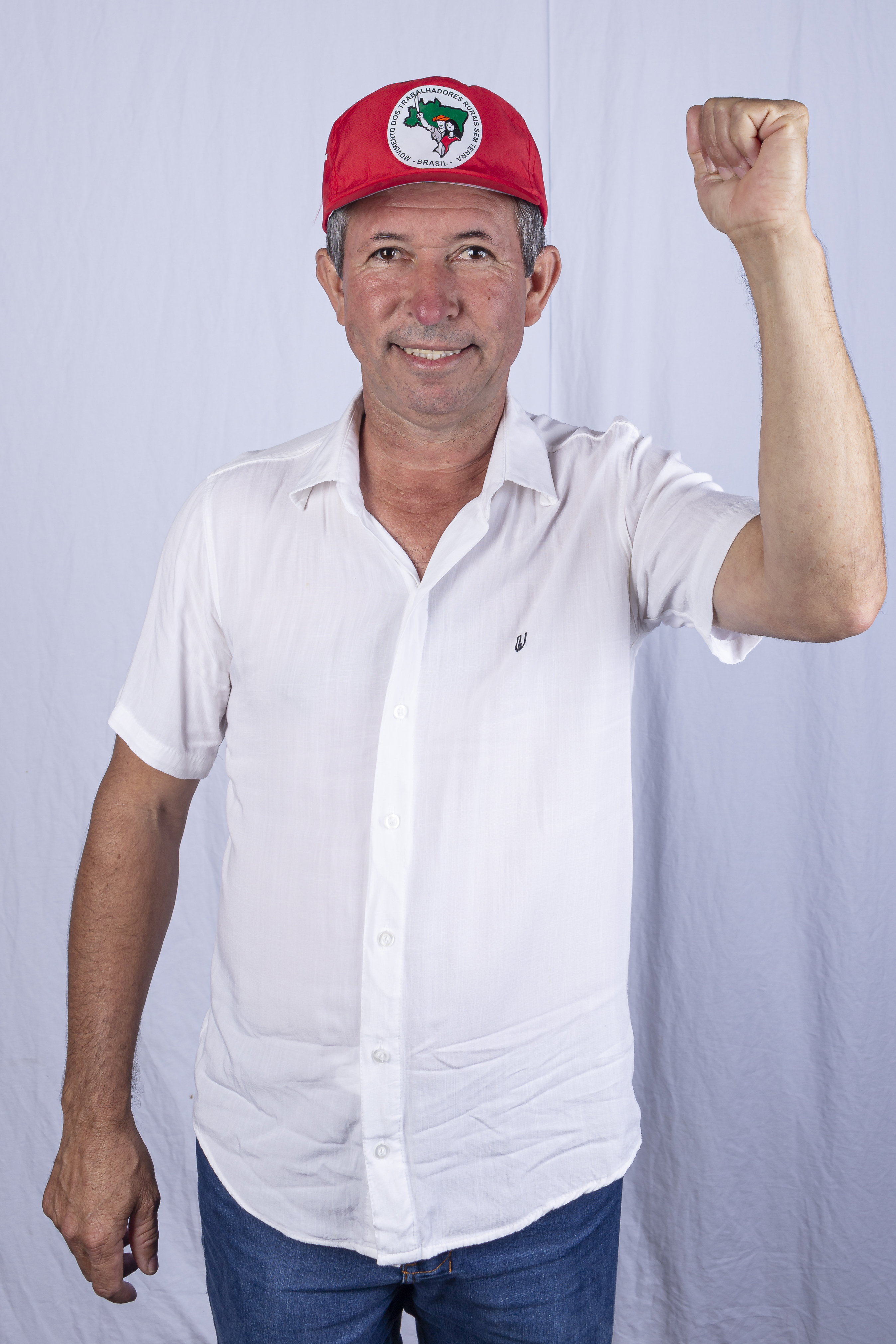 Foto de Edilson Sem Terra, homem idoso, branco, de cabelos curtos grisalhos. Ele está sorrindo e olhando fixamente para a câmera. Ele usa uma camisa branca de botões, calça jeans e está com o punho fechado e levantado.