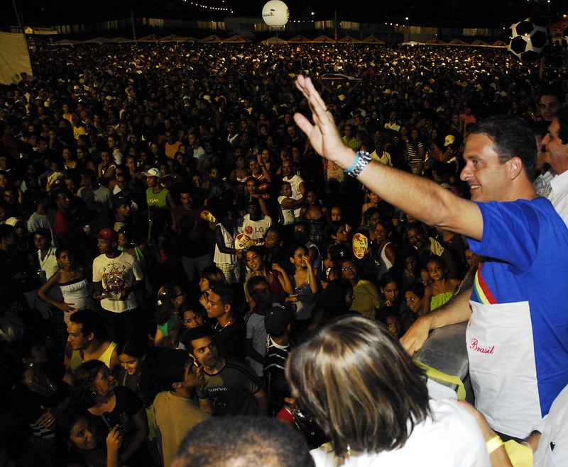 Foto noturna de Eduardo campos saudando multidão. Ele está em um palanque acenando com a mão esquerda, usando uma camisa com as cores azul e branco da bandeira de Pernambuco.