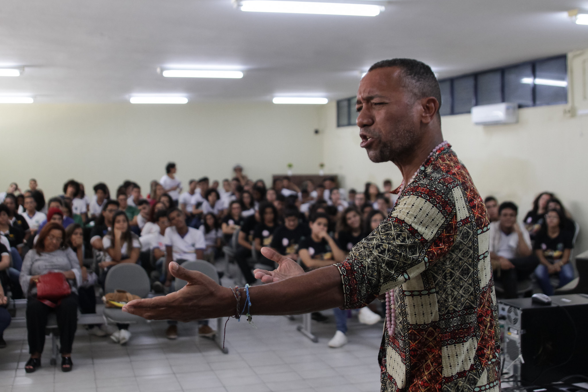 Foto de Miró da Muribeca se apresentando diante de uma plateia que lota um auditório bem iluminado, com paredes e pisos brancos. Ele é um homem negro de meia idade e cabelos curtos vestindo uma camisa com estampas geométricas nos tons cinza, marrom e creme. A plateia, apesar de desfocada, parece ser formada por pessoas jovens.