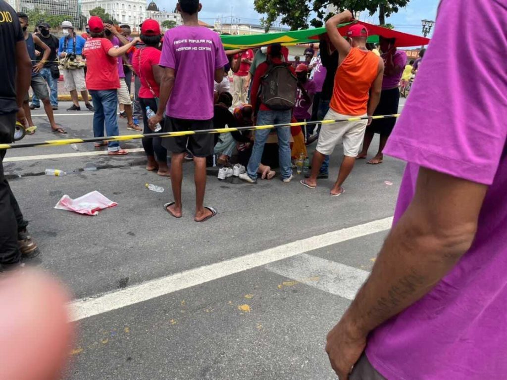 A imagem mostra um grupo de pessoas reunidas ao ar livre, participando de um evento público ou manifestação. Há uma fita amarela que parece estar delimitando uma área para possibilitar o atendimento à advogada atropelada, que não aparece na imagem por estar no chão, cercada pelas pessoas que prestam socorro. As pessoas mais próximas da câmera estão vestindo camisetas roxas, e há outras ao fundo com camisetas vermelhas e algumas segurando um bandeira do Brasil para fazer sombra sobre a mulher atropelada. O cenário parece ser uma área urbana, com estradas e grama visíveis ao fundo.