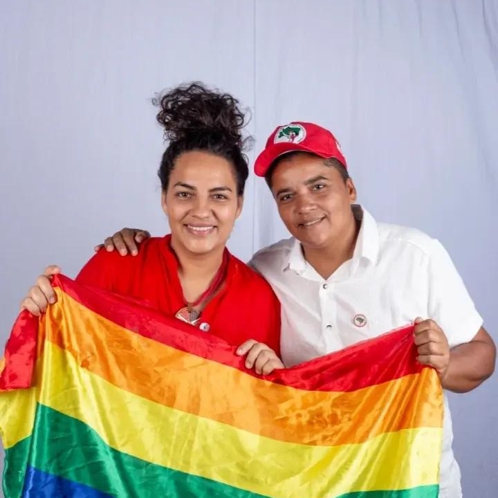 Foto de Rosa Amorim e Zezinha do MST. As duas são mulheres jovens, negras e estão segurando uma bandeira do arco-íris simbolizando o movimento LGBT. Rosa está à esquerda, usando uma camiseta vermelha, com o cabelo preto preso em coque no alto da cabeça. Zezinha tem cabelos curtos, ocultos pelo boné vermelho do MST e usa uma camisa branca de botões.