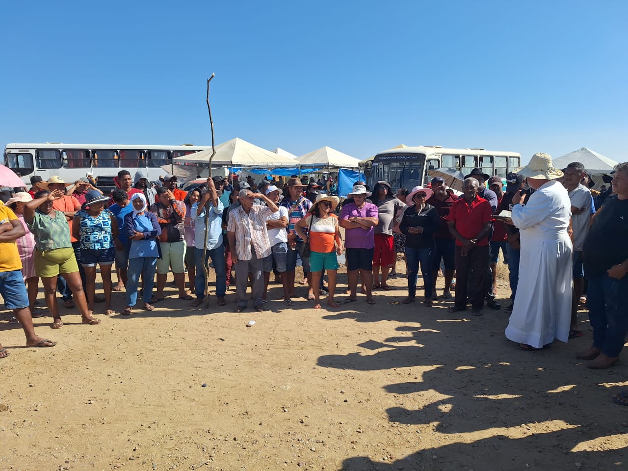 A imagem mostra um grande grupo de pessoas ao ar livre, alinhadas, com as mãos cobrindo os olhos, como se estivessem protegendo-os do sol. Atrás do grupo, há várias tendas brancas e ônibus estacionados em uma área que parece ser um campo seco e arenoso, sob um céu azul claro. As pessoas estão vestindo roupas e chapéus de várias cores, sugerindo que pode ser um dia quente e ensolarado.