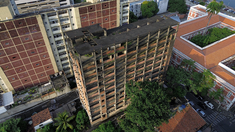 A imagem mostra uma vista aérea de um prédio alto e desgastado, cercado por outras construções. O prédio em destaque tem uma fachada laranja desbotada e está densamente coberto por vegetação, indicando possível abandono ou falta de manutenção. Ao lado dele, há edifícios com telhados de telha vermelha, e uma rua é visível ao lado das construções. A presença de palmeiras sugere um clima quente. A imagem é interessante devido ao contraste entre o prédio coberto de vegetação e as estruturas mais bem cuidadas ao redor,