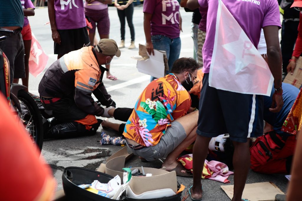 A imagem mostra uma cena ao ar livre onde uma pessoa está recebendo atendimento médico na rua. Há várias pessoas ao redor, algumas vestindo camisetas roxas e outras com roupas diferentes. Duas pessoas com coletes laranja, provavelmente paramédicos ou socorristas, estão atendendo a pessoa no chão, que parece estar sentada. Há suprimentos médicos visíveis no chão ao lado de uma bolsa preta, sugerindo que um tratamento médico está sendo administrado.