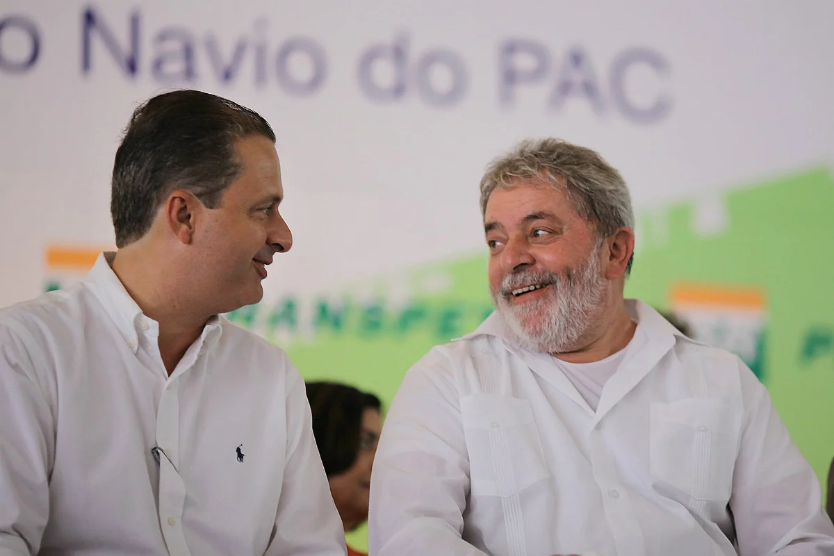 Foto colorida de Eduardo Campos, à esquerda, e Lula, à direita da imagem. Ambos estão de camisa branca e estão sentados diante de um painel branco com detalhes verdes onde é possível se ler Navio do PAC. Os dois estão sorrindo em meio a uma conversa descontraída.