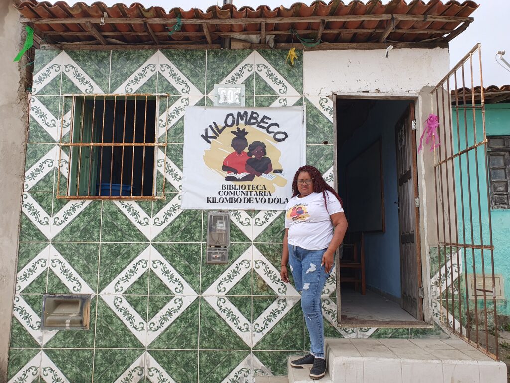 A imagem mostra a fachada de uma pequena casa com uma placa que diz “KILOMBECO BIBLIOTECA COMUNITÁRIA KILOMBO DE VÓ DÔLA”. A casa tem uma fachada de azulejos com padrões geométricos verdes e brancos. À esquerda, há uma janela com grades e, à direita, uma porta aberta que leva para o interior. Junto da grade da porta aberta há uma mulher jovem, negra, de óculos, cabelos compridos por cima dos ombros vestindo roupas casuais (camiseta branca e calça jeans). Ela está de pé encostada à parede de azulejos.