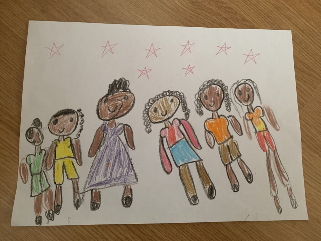 A imagem é um desenho infantil que mostra seis figuras que representam pessoas negras, desenhadas com giz de cera ou lápis de cor. As figuras estão lado a lado e variam em cores, sugerindo diversidade. Cada figura tem diferentes estilos de cabelo e cores de roupas, como verde, amarelo, roxo, rosa, laranja e marrom. Acima das figuras, há estrelas desenhadas em tons de vermelho e rosa. O fundo é uma folha de papel branca colocada sobre uma superfície de madeira.