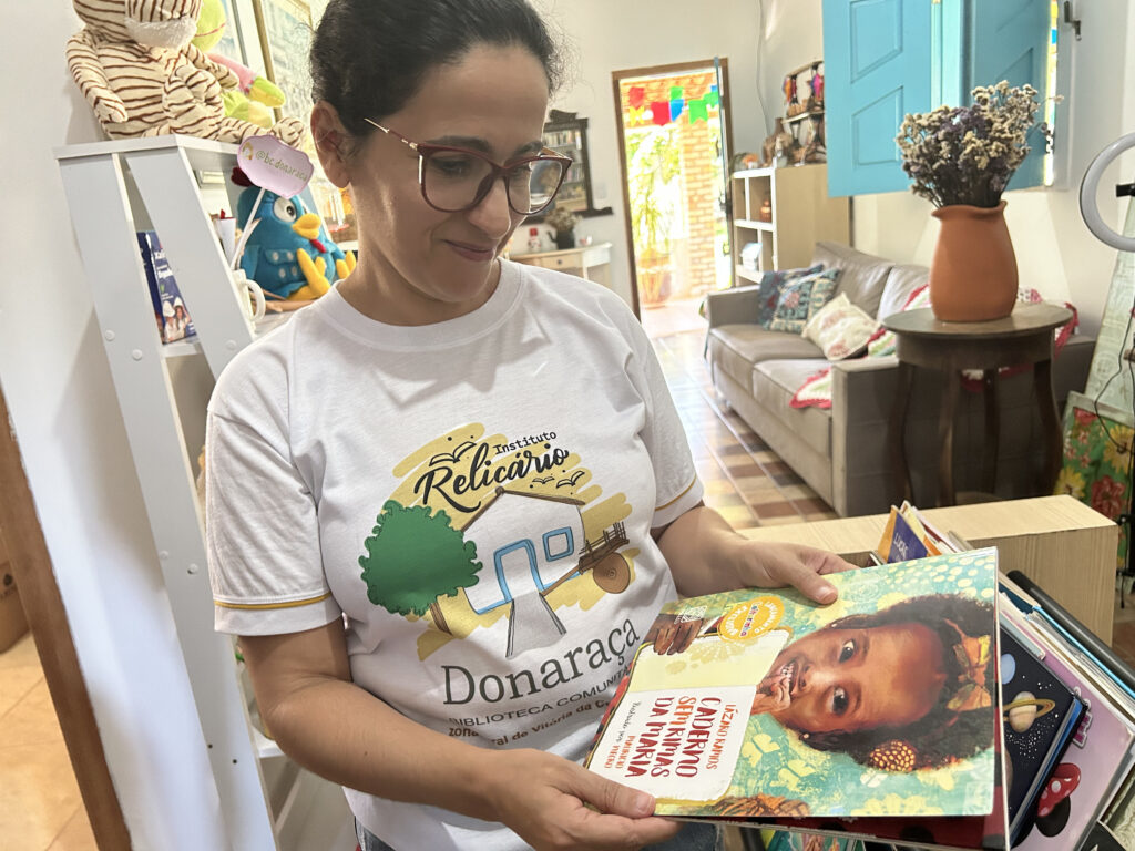 A foto mostra uma mulher de pele clara, óculos de aros grandes, com cabelos pretos presos por trás do pescoço, em um ambiente interno, segurando uma pilha de livros coloridos. A pessoa está vestindo uma camiseta branca com desenhos gráficos e texto. No fundo, há vários objetos, incluindo um modelo de esqueleto à esquerda, uma porta azul no centro e itens decorativos como vasos e quadros na parede.