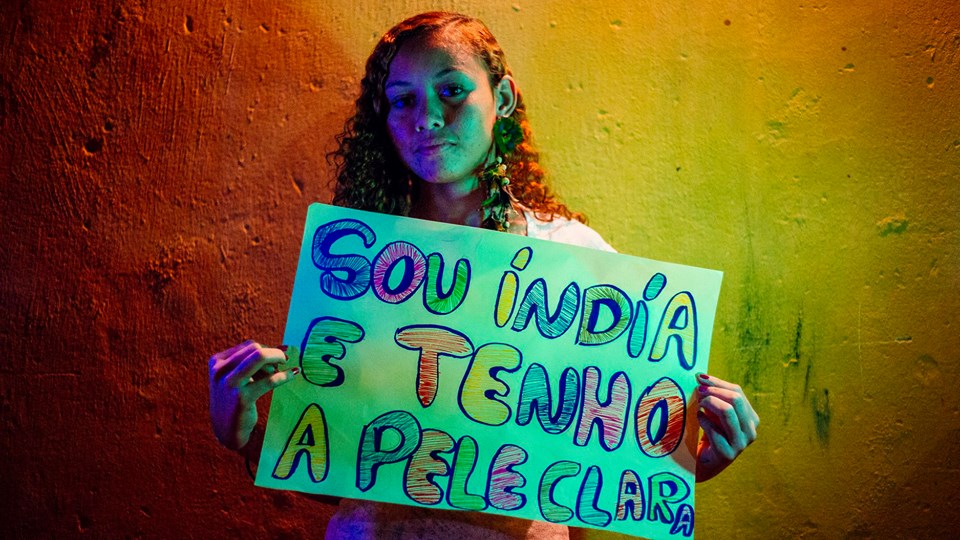 A imagem mostra uma mulher jovem em pé contra um fundo colorido com tons de amarelo e verde. A moça tem cabelos compridos ondulados e olha direto para a câmera enquanto segura um cartaz com a mensagem escrita à mão, com letras coloridas, em português: “Sou indígena e tenho a pele clara.”