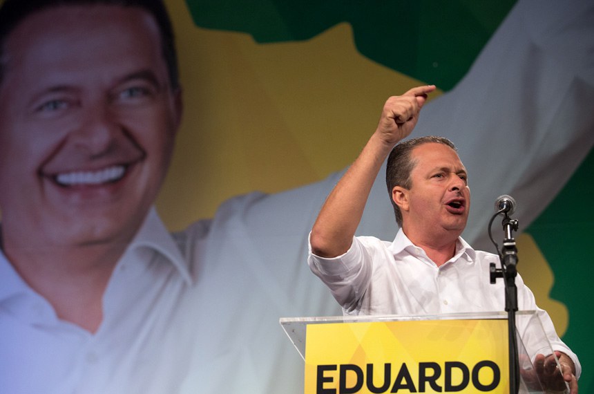 Foto de Eduardo Campos, homem branco, com cabelos claros, bem penteados e com calvície acentuada, discursando diante de um microfone colocado sobre um púlpito onde se lê o nome Eduardo em letras pretas sobre fundo amarelo. Ao fundo, há outra foto do próprio Eduardo Campos, em um grande banner por trás do orador.