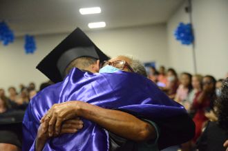 A imagem retrata um close de duas pessoas se abraçando. Uma delas está vestindo uma beca e um capelo de formatura, indicando que se trata de uma cerimônia de graduação. A beca é roxa, e o capelo tem um formato quadrado e plano. A outra pessoa parece ser mais velha, com cabelos grisalhos e óculos. Ambas estão com os braços entrelaçados em um abraço. Infelizmente, não conseguimos ver os rostos delas devido ao ângulo da foto e à posição do abraço. Ao fundo, há figuras borradas que podem ser outros participantes da cerimônia, sugerindo que esse momento está sendo compartilhado em meio a uma celebração maior.