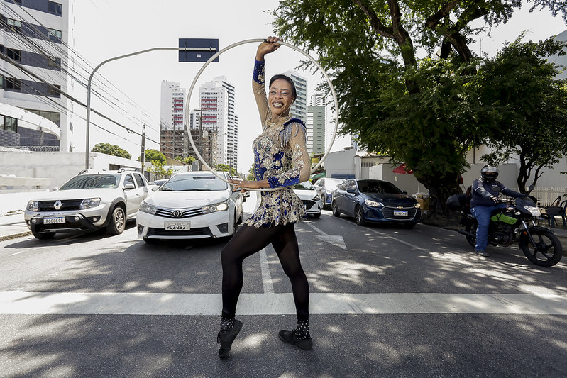 A imagem mostra uma mulher negra e jovem no meio de uma rua, aparentemente realizando uma dança ou performance com um arco (bambolê) de ginástica rítmica. Ela está vestindo um traje elaborado com elementos brilhantes. O traje inclui um adereço de cabeça e é predominantemente azul com estampas coloridas prata e douradas. A pose da mulher envolve um braço levantado acima da cabeça e o outro ao lado, sugerindo movimento. Ao redor do performer, há carros e motocicletas, indicando que a cena ocorre em uma rua ativa, no meio do tráfego. Prédios alinham a rua ao fundo, sugerindo um ambiente urbano.